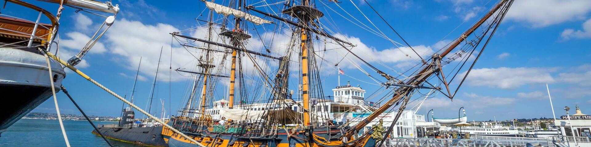 Un gran barco de vela atracado en un puerto con cielo azul despejado y un paisaje urbano al fondo.
