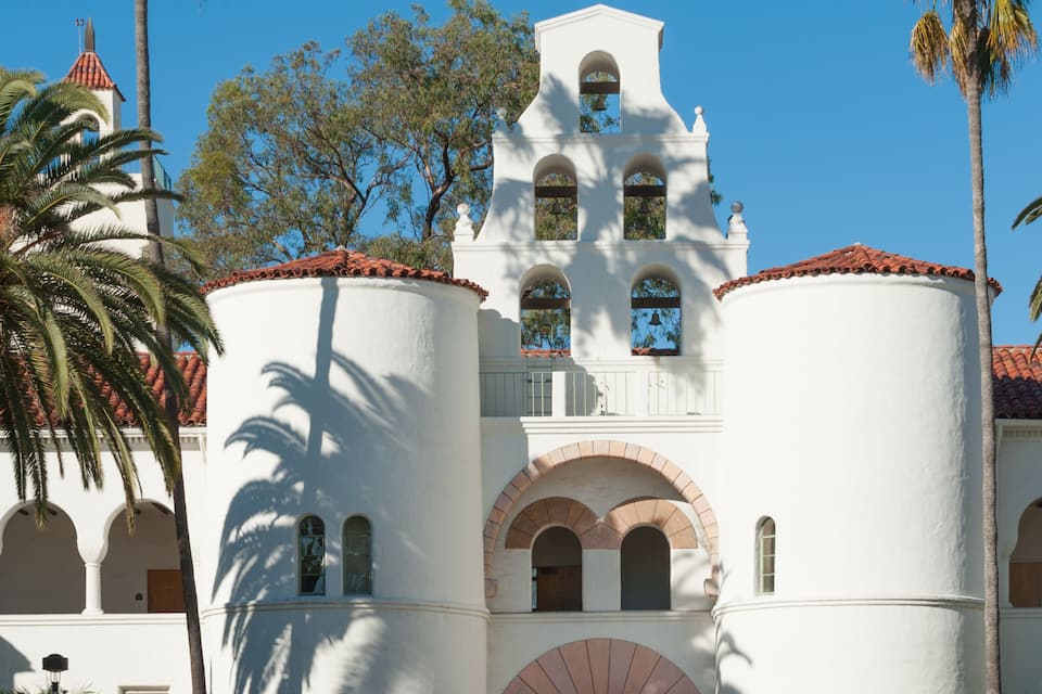 A white building with arched windows and a bell tower, surrounded by palm trees and a circular flower bed.