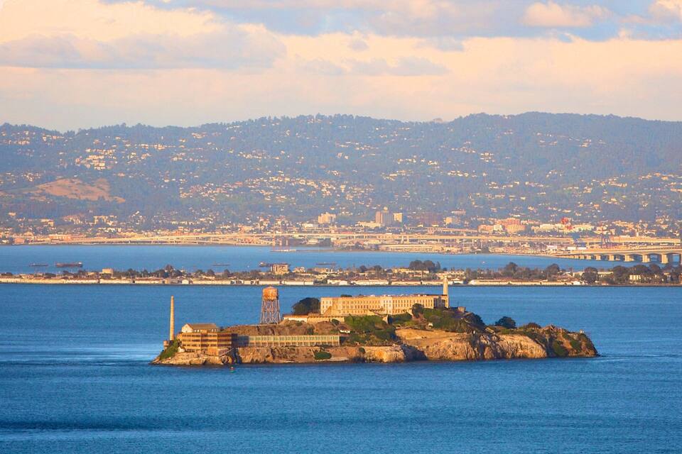 Una città costiera con una fortezza isolana ben visibile, un ponte e uno sfondo collinare.