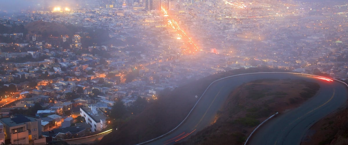 A cityscape at dusk with a winding road and a bridge in the distance.