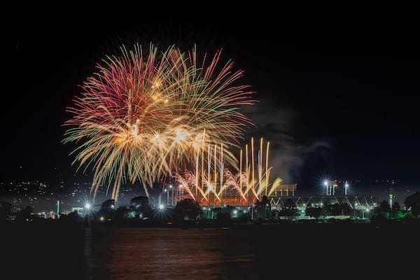 A nighttime fireworks display over a cityscape with illuminated buildings and a body of water in the foreground.