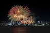 Un espectáculo de fuegos artificiales nocturnos sobre un paisaje urbano con edificios iluminados y una masa de agua al frente.