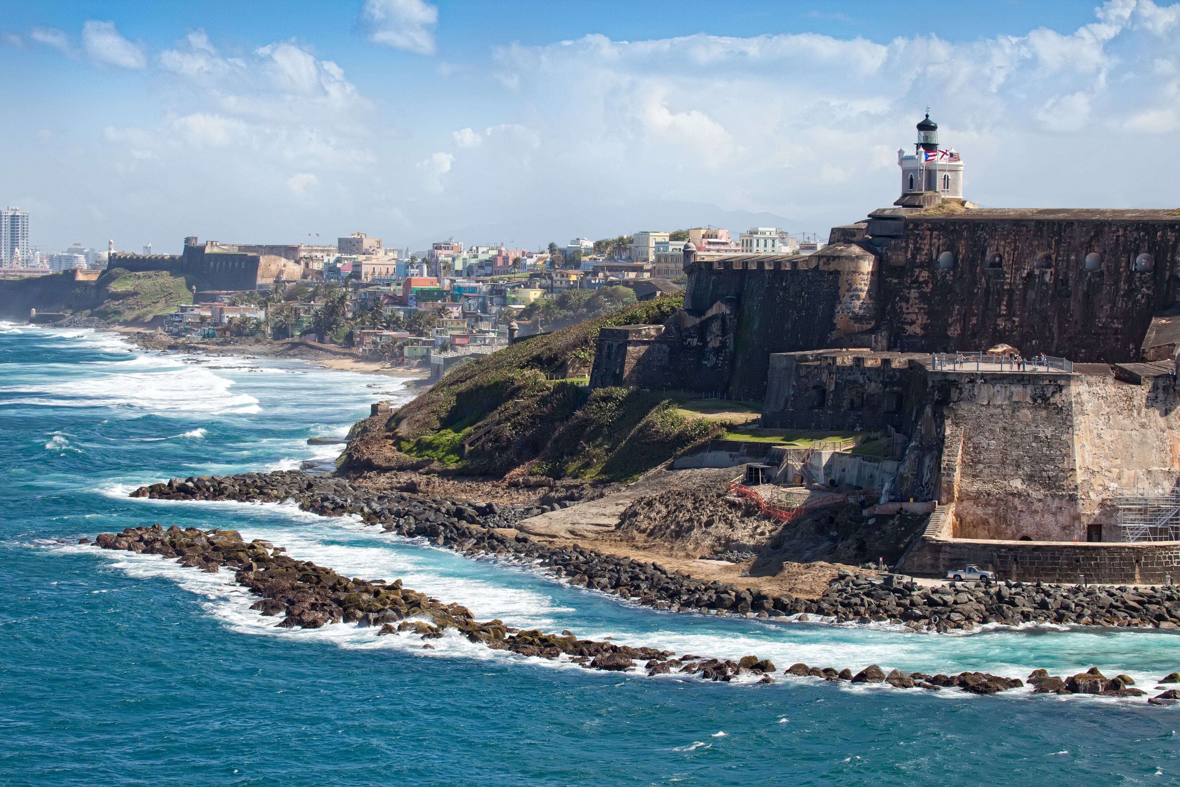 Uma fortificação costeira com um farol, litoral rochoso e um horizonte urbano ao fundo.