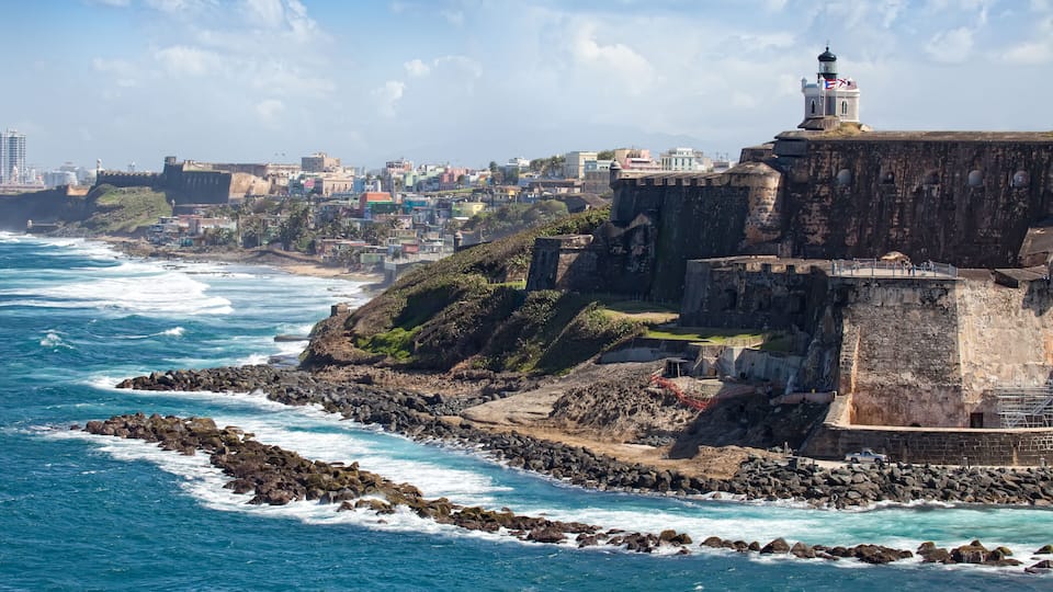 Uma fortificação costeira com um farol, litoral rochoso e um horizonte urbano ao fundo.
