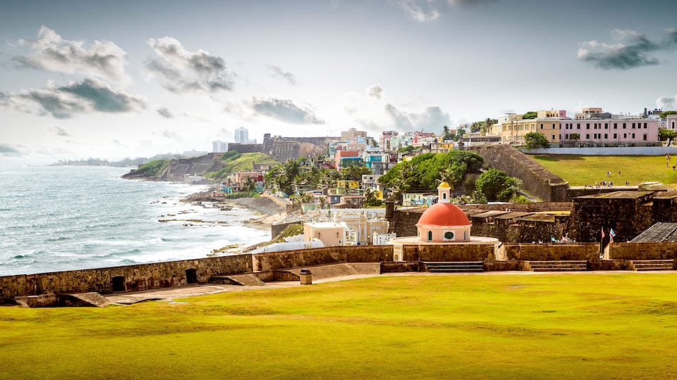 A coastal cityscape with a prominent red-domed building, historic fortifications, and a mix of colorful residential buildings along the shore.