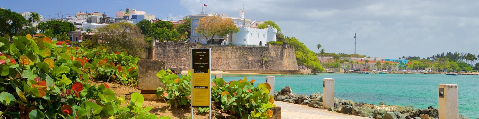 Um calçadão costeiro com uma placa, vegetação exuberante e uma fortaleza histórica com vista para o mar.