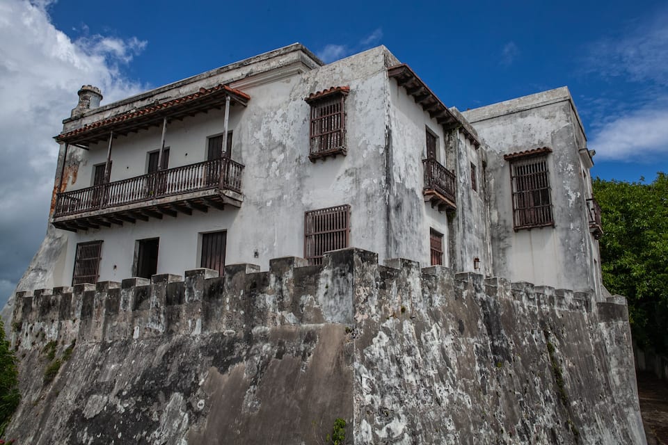 A weathered building with a balcony and barred windows, surrounded by a high stone wall.