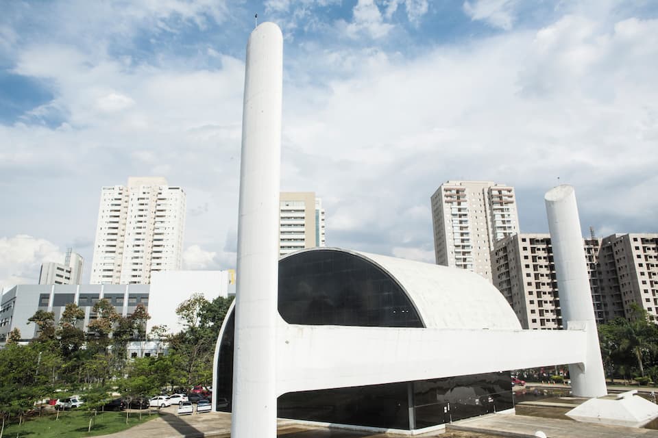 Una struttura architettonica moderna con un tetto curvo, circondata da alti edifici residenziali e un cielo sereno.