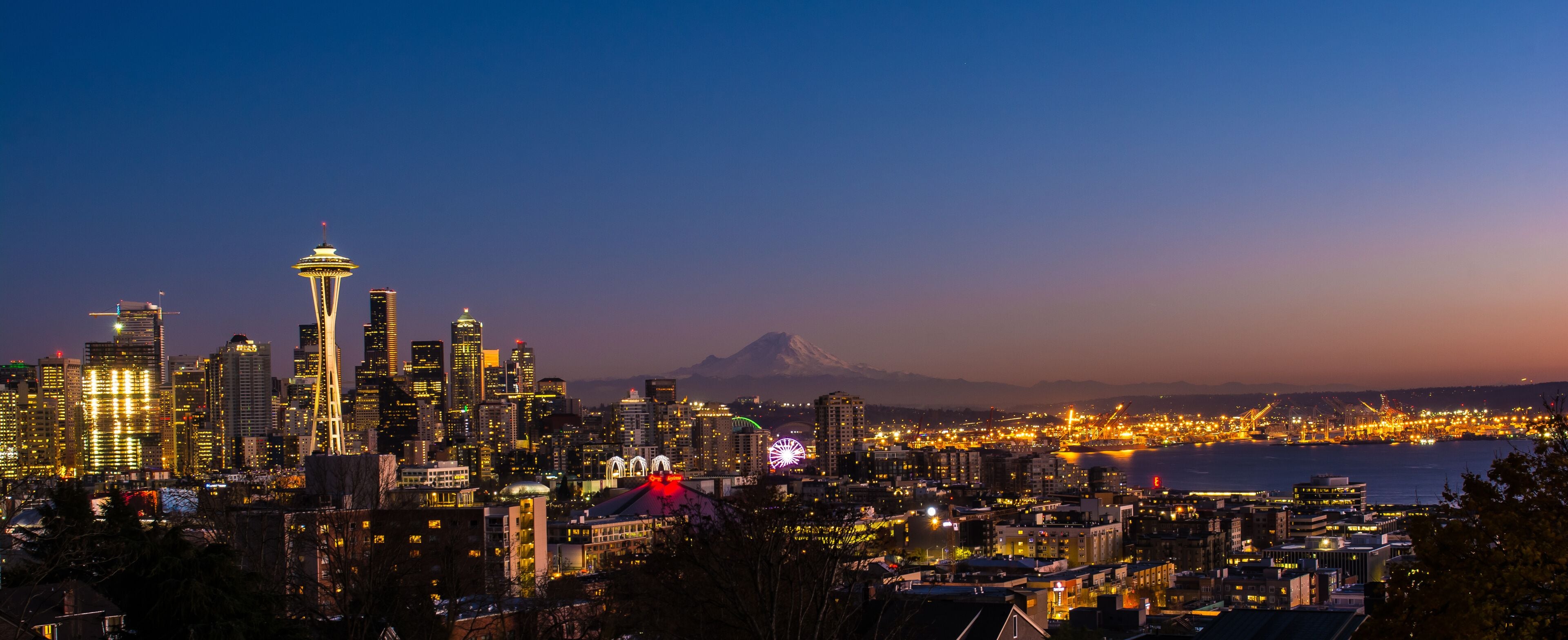 A city skyline at dusk with illuminated buildings, a prominent tower, and a mountain in the background.