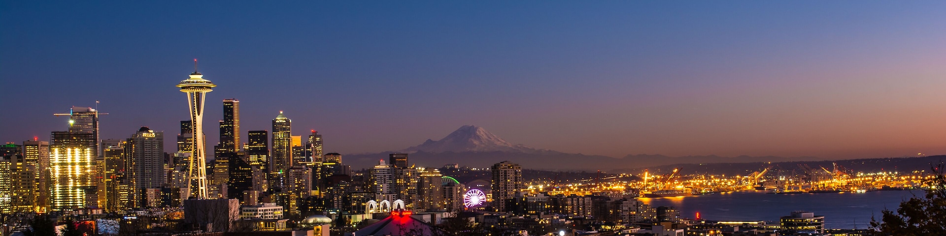 A city skyline at dusk with illuminated buildings, a prominent tower, and a mountain in the background.