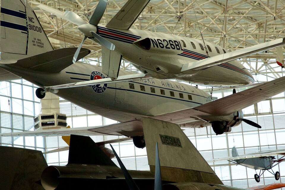 Une collection d’aéronefs, dont un avion blanc portant les inscriptions « Alaska Airlines » et « NC 91008 », est exposée dans un grand hangar ouvert.