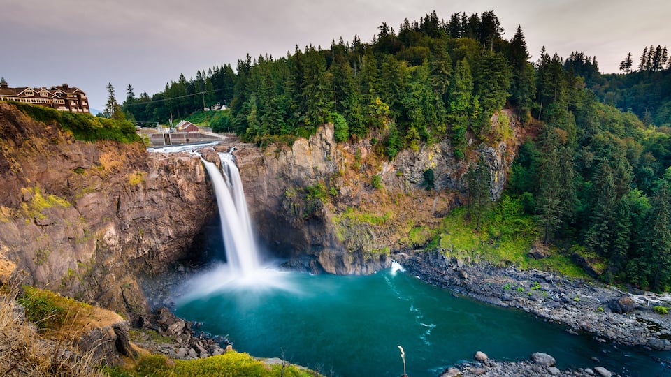 A waterfall cascading into a turquoise pool surrounded by forested hills and a lodge.