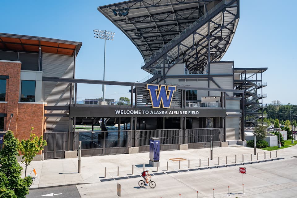 A modern stadium with a large 'W' logo, a sign welcoming visitors to Alaska Airlines Field, and a cyclist on the sidewalk.