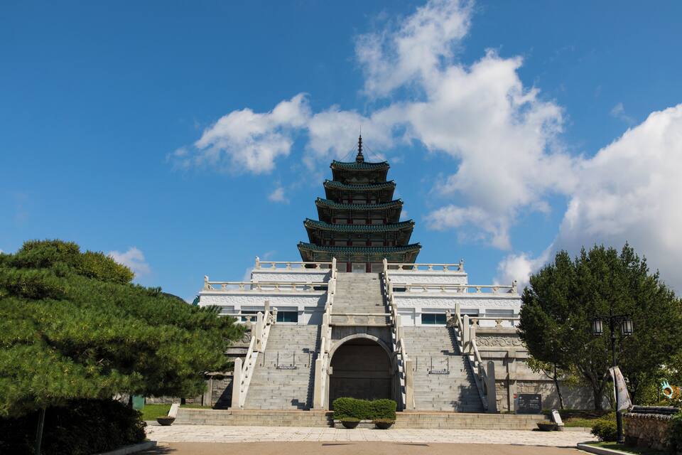 A traditional multi-tiered pagoda with a staircase leading up to it, surrounded by greenery and a clear blue sky.