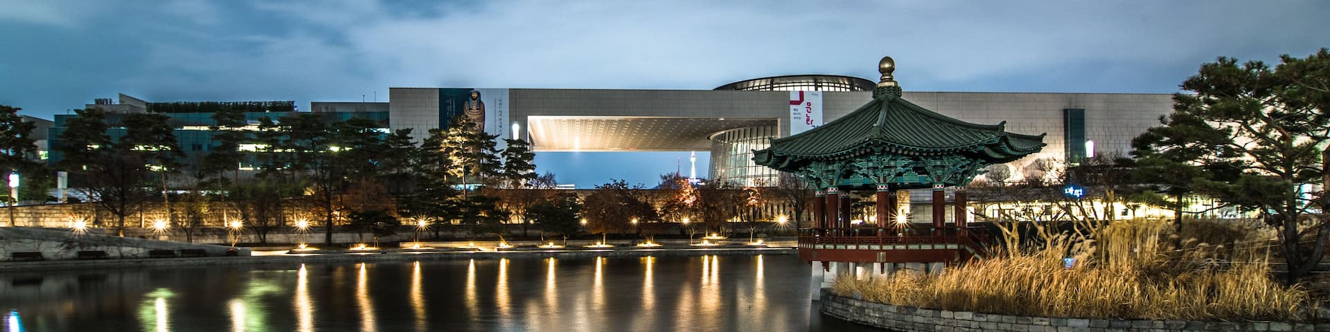 A nighttime view of a modern building with a traditional pavilion by a calm water body.