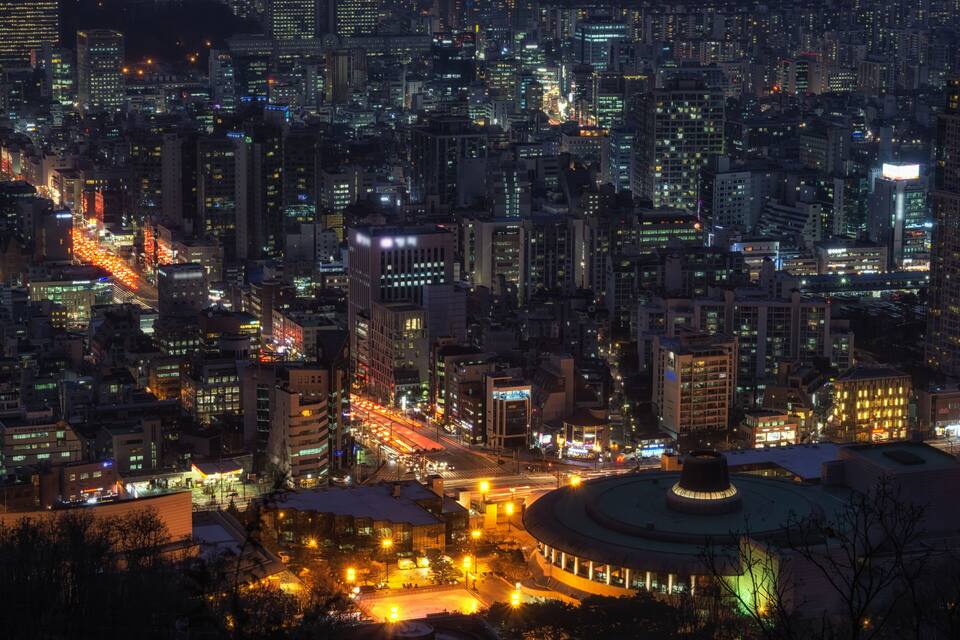 Un paisaje urbano nocturno con edificios densos, calles iluminadas y un edificio circular destacado con cúpula.