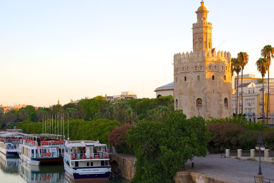 Una torre histórica con un minarete, barcos amarrados a lo largo de un río y abundante vegetación.
