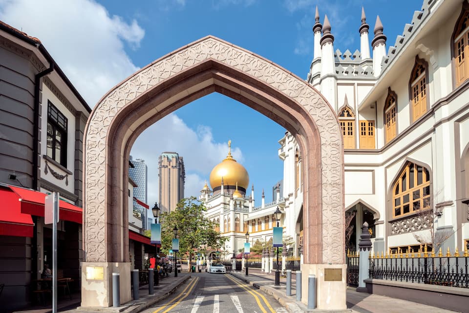 A street with a large archway, a golden dome, and white buildings with pointed windows.
