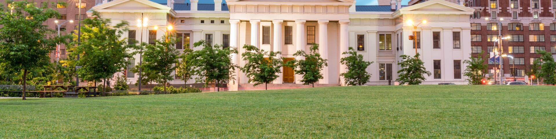 A large domed building with a prominent arch structure in front, surrounded by modern buildings and a grassy area.