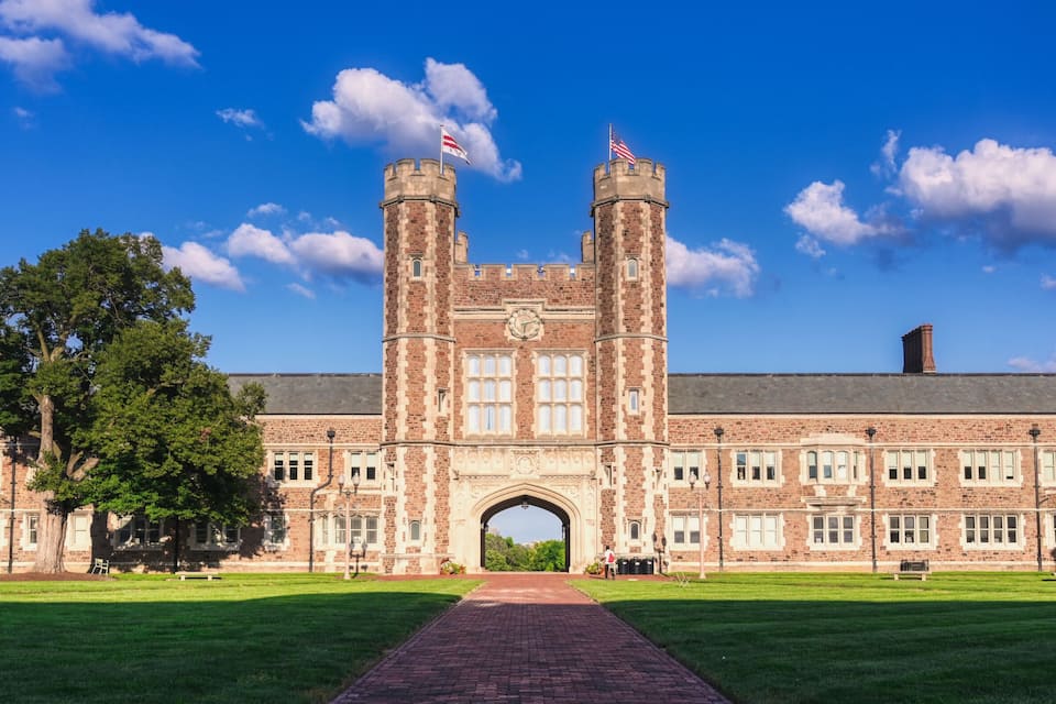 A large brick building with a central archway, two towers, and a flag.