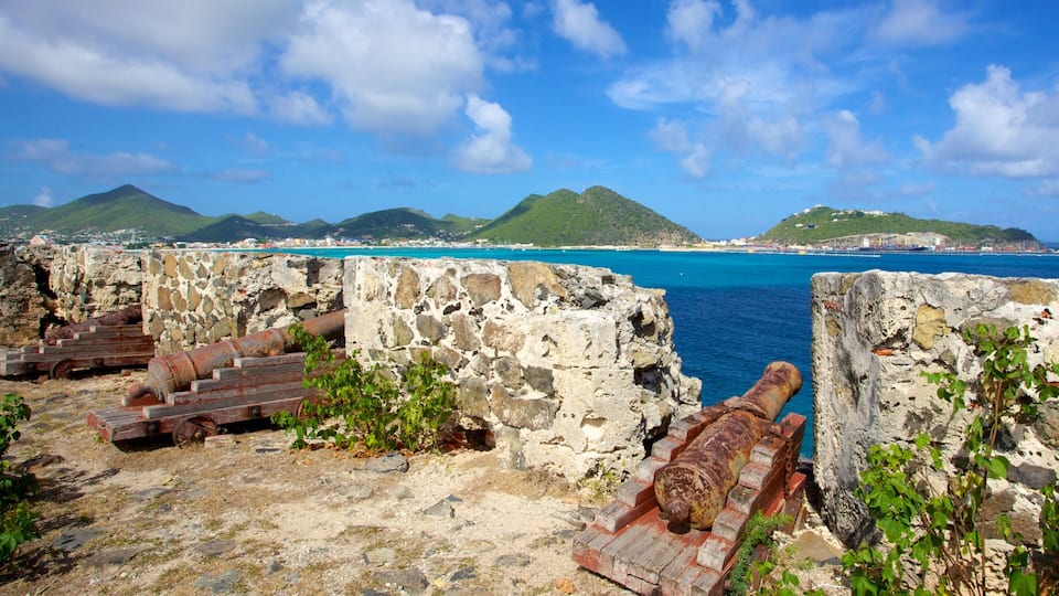 A coastal fortification with rusted cannons, a clear blue sea, and green hills in the background.