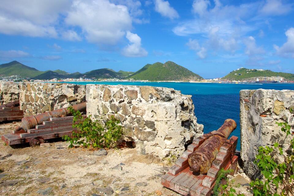 A coastal fortification with rusted cannons, a clear blue sea, and green hills in the background.