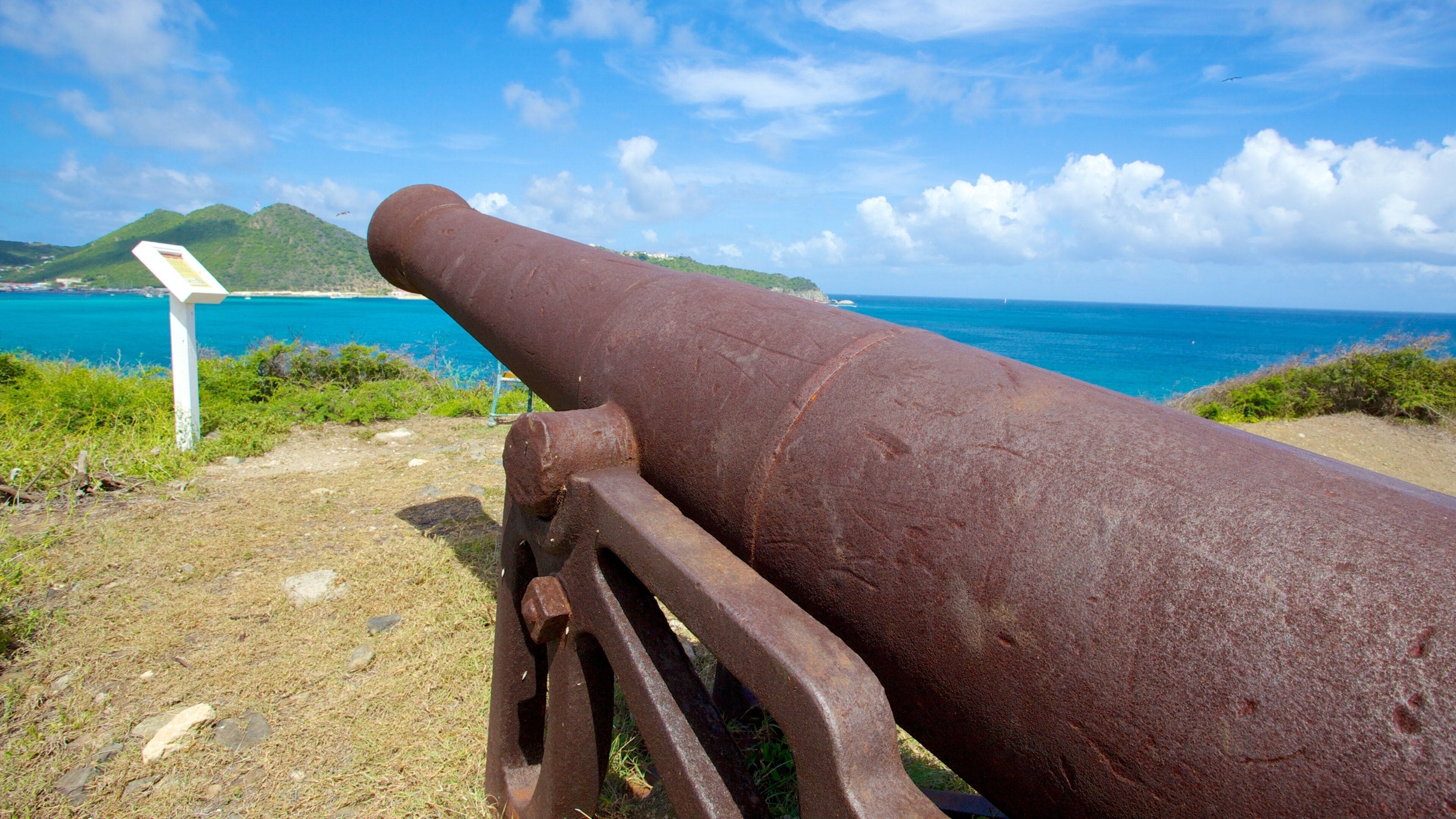 A rusted cannon overlooking a coastal landscape with a clear sky and a signpost in the foreground.