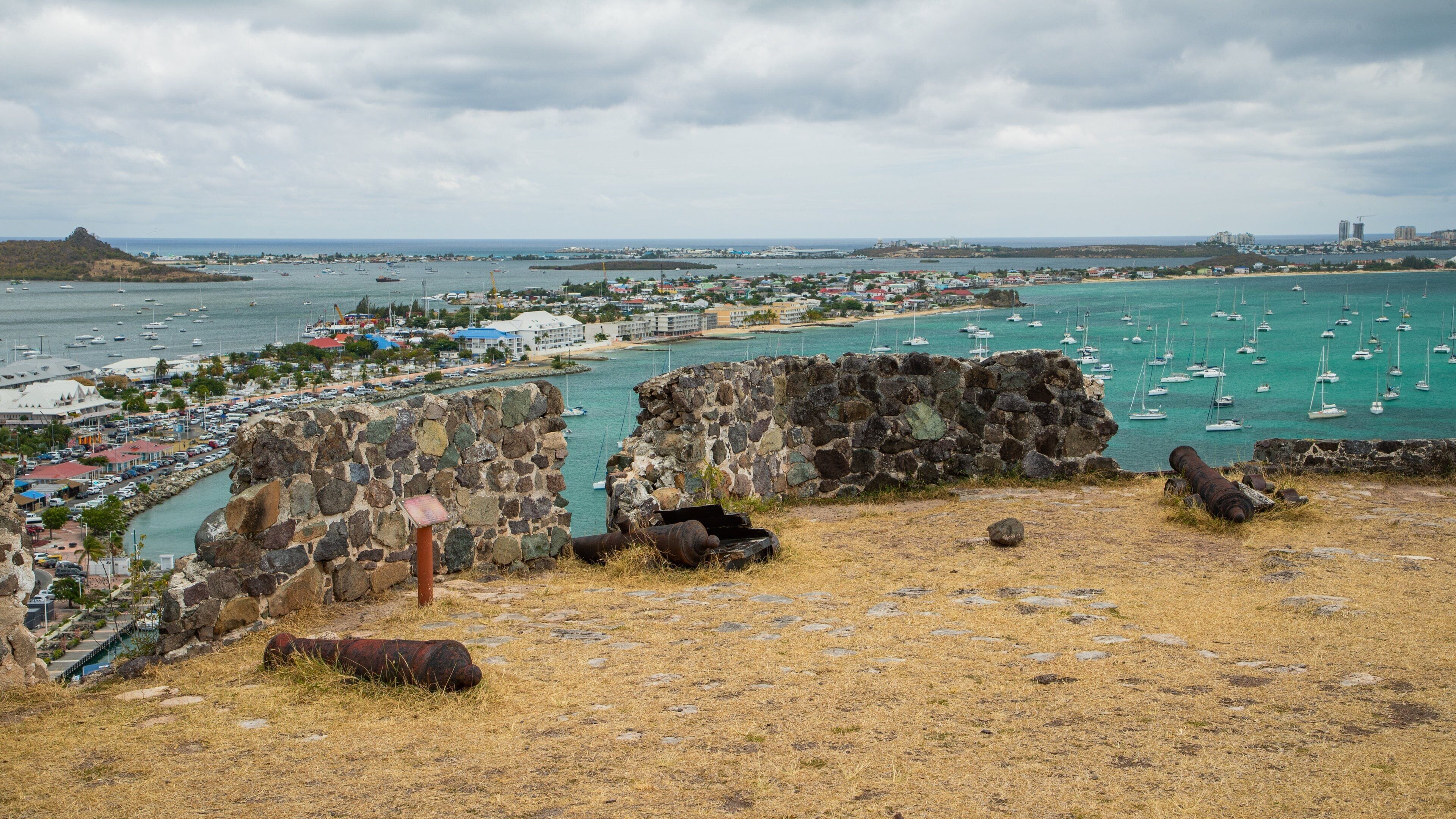 Un pueblo costero con un fuerte histórico, un puerto deportivo lleno de barcos y una montaña a lo lejos.