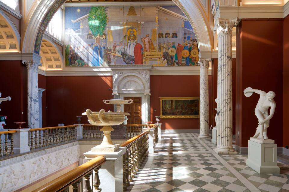 A grand hall with a checkered floor, classical columns, a large fresco, and a statue.