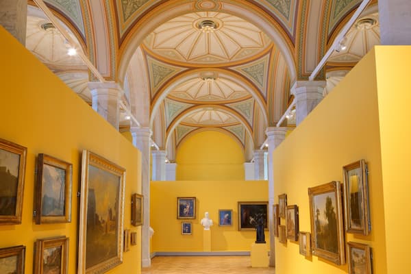 A gallery with yellow walls and a high ceiling with intricate designs.