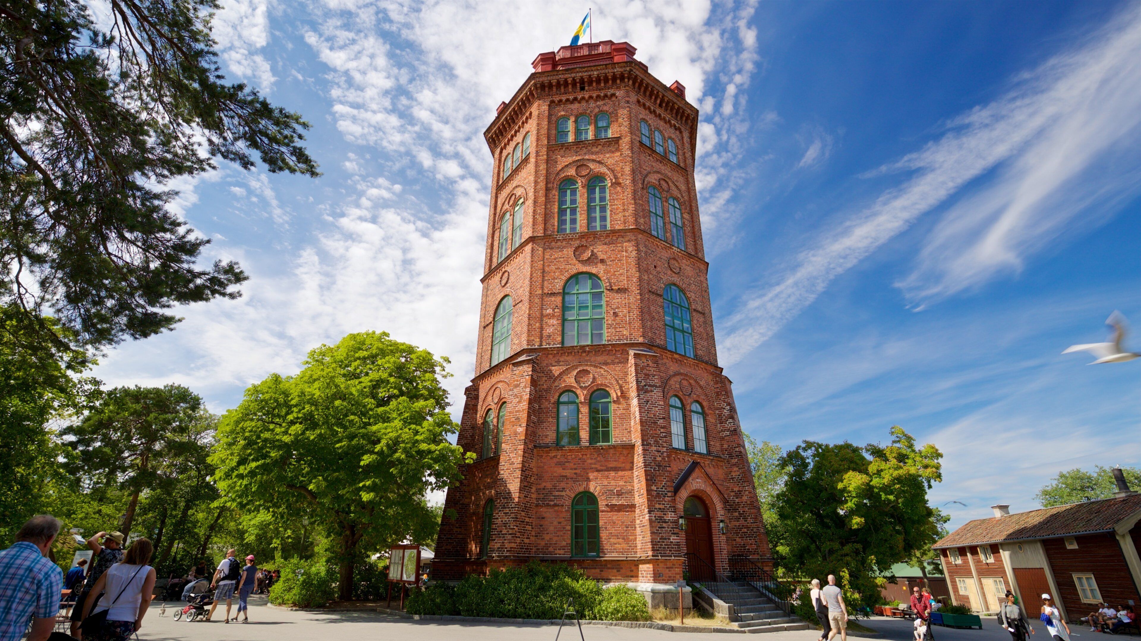 A tall brick tower with green windows and a flag on top, surrounded by trees and people.