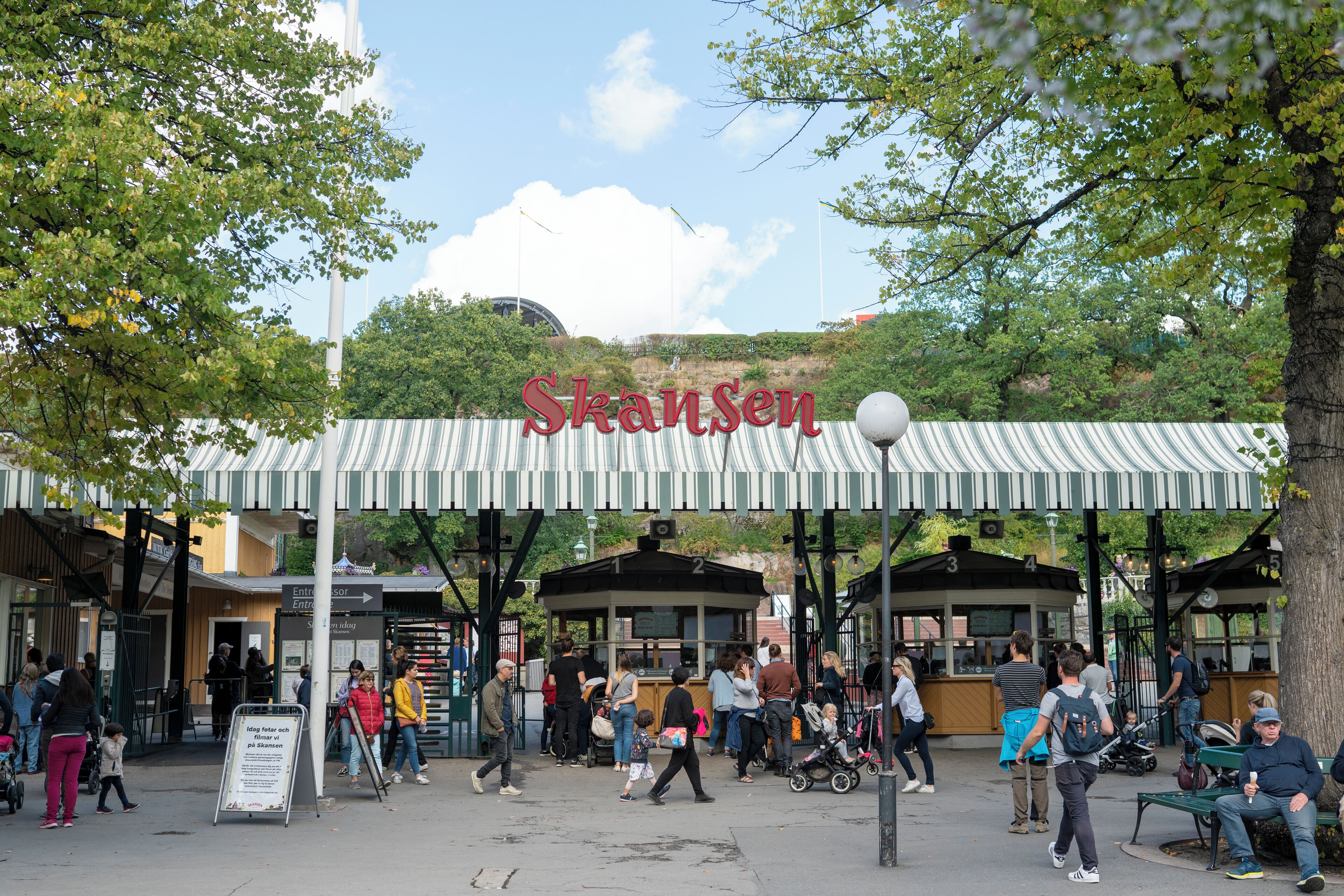 Vilkkaalla ulkoilmamarkkinalueella on raidallinen markiisi, jossa lukee ”Skansen”, ja siellä on useita ihmisiä kävelemässä ympäriinsä.