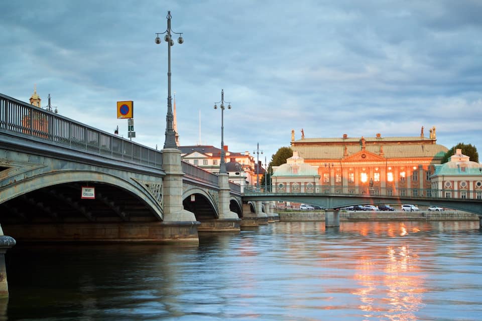 A bridge over a river with a reflection of lights on the water. There is a building with a dome and a sign that says "INFART FÖRBIUDEN" on the bridge.