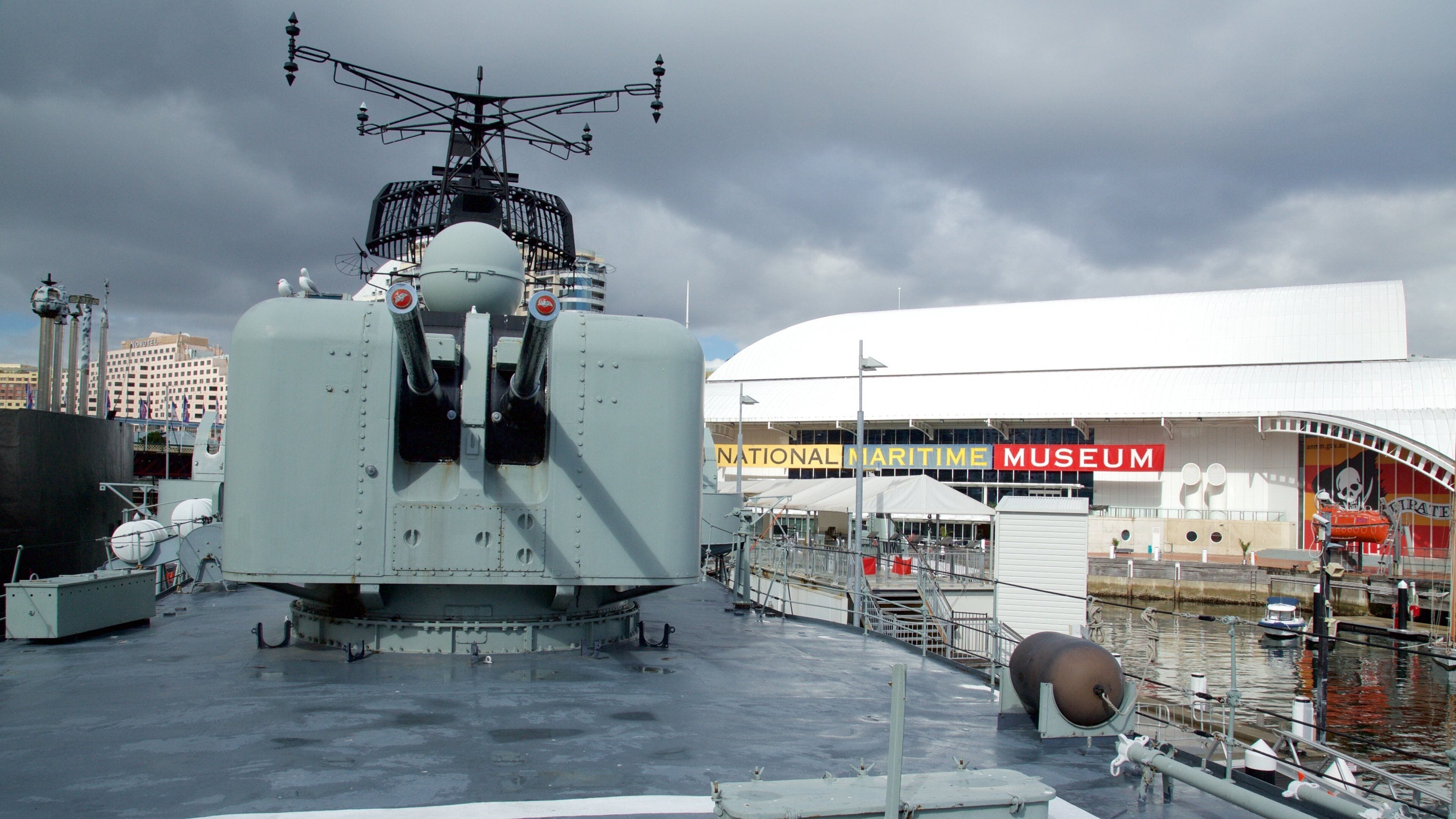 A large military ship docked at a pier with the National Maritime Museum in the background.
