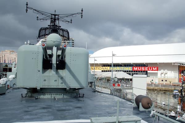 A large military ship docked at a pier with the National Maritime Museum in the background.