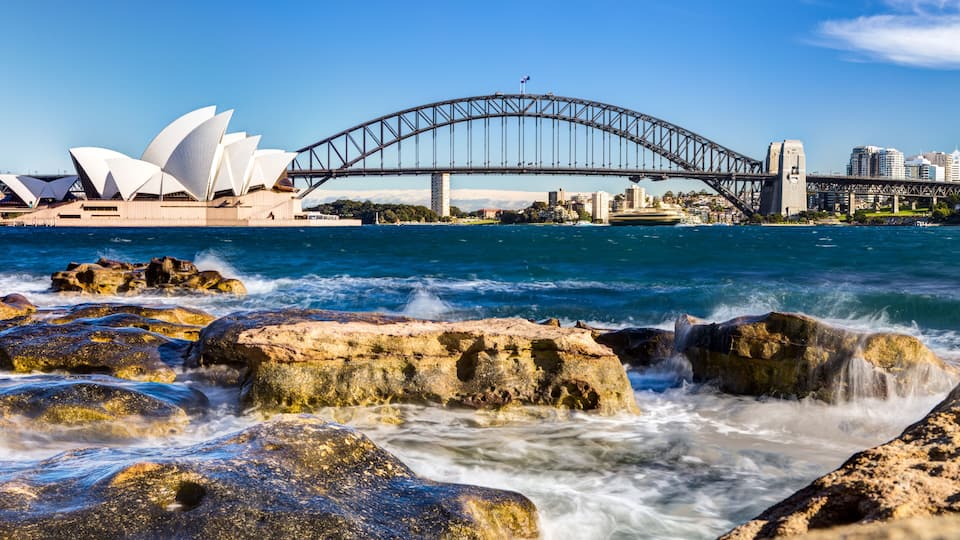 La Casa de la Ópera de Sydney y el Puente de Sydney con una costa rocosa en primer plano.