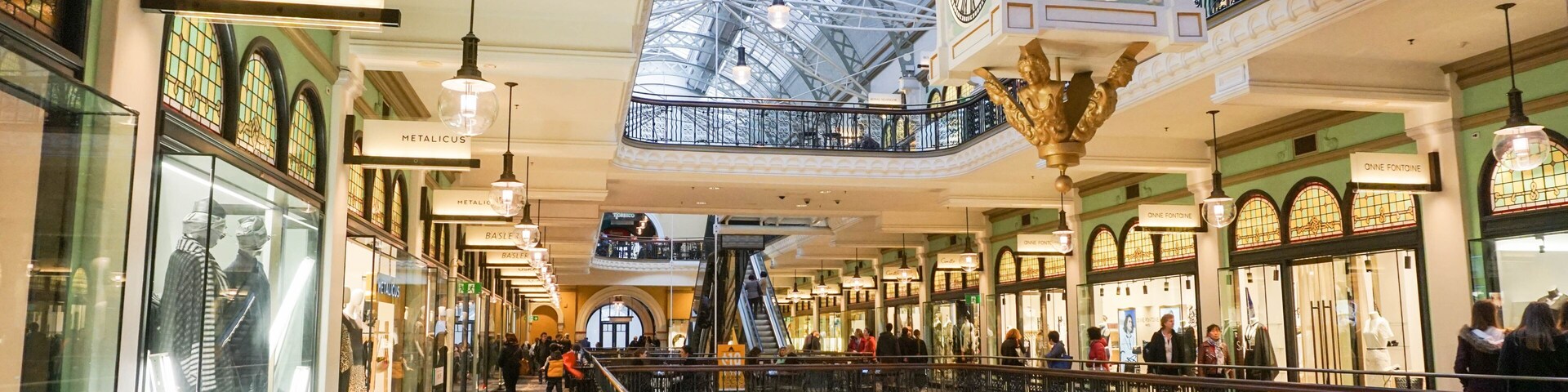 Um shopping center com teto de vidro, grades ornamentadas e vários andares.