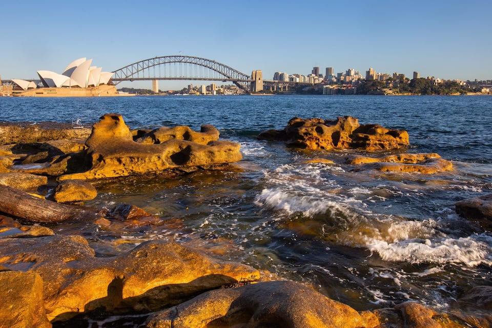 Rocky coastline with waves, Sydney Opera House, and Sydney Harbour Bridge.