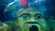 An underwater scene with a large, textured sculpture resembling a face, surrounded by fish and a shark in the background.