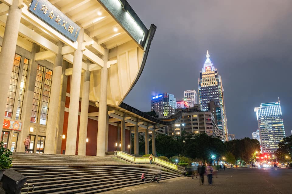 A grand building with large columns and a sign in a foreign language. In the background, there are illuminated skyscrapers and a busy street with people walking.