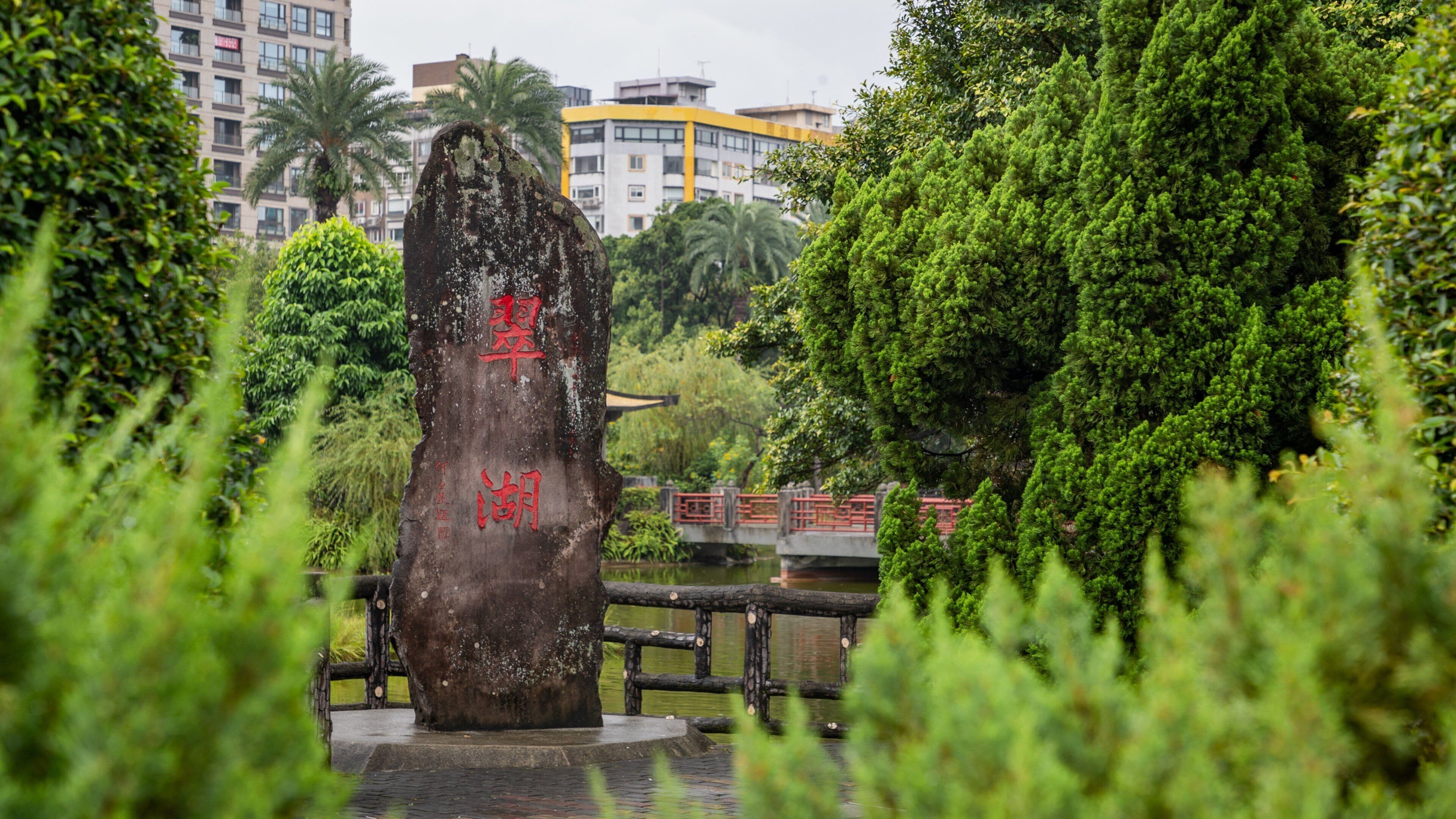 A stone monument with red inscriptions, surrounded by lush greenery and a bridge in the background.