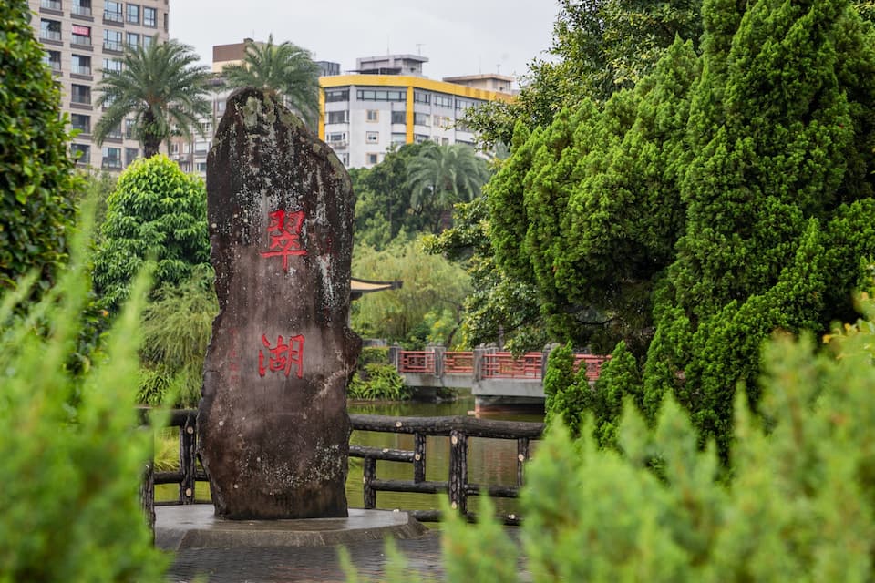 A stone monument with red inscriptions, surrounded by lush greenery and a bridge in the background.