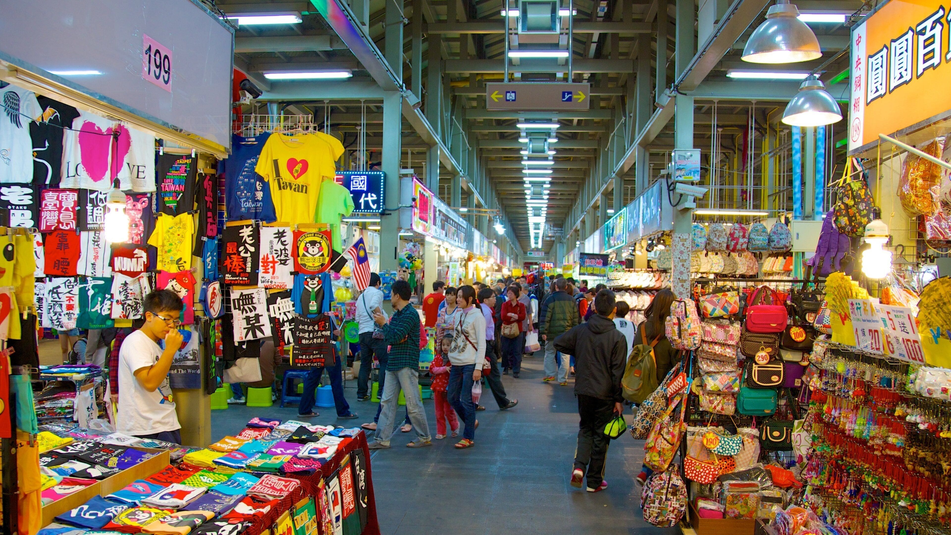 Un marché intérieur animé, avec de nombreux étals proposant une variété de produits colorés, notamment des sacs et des vêtements.