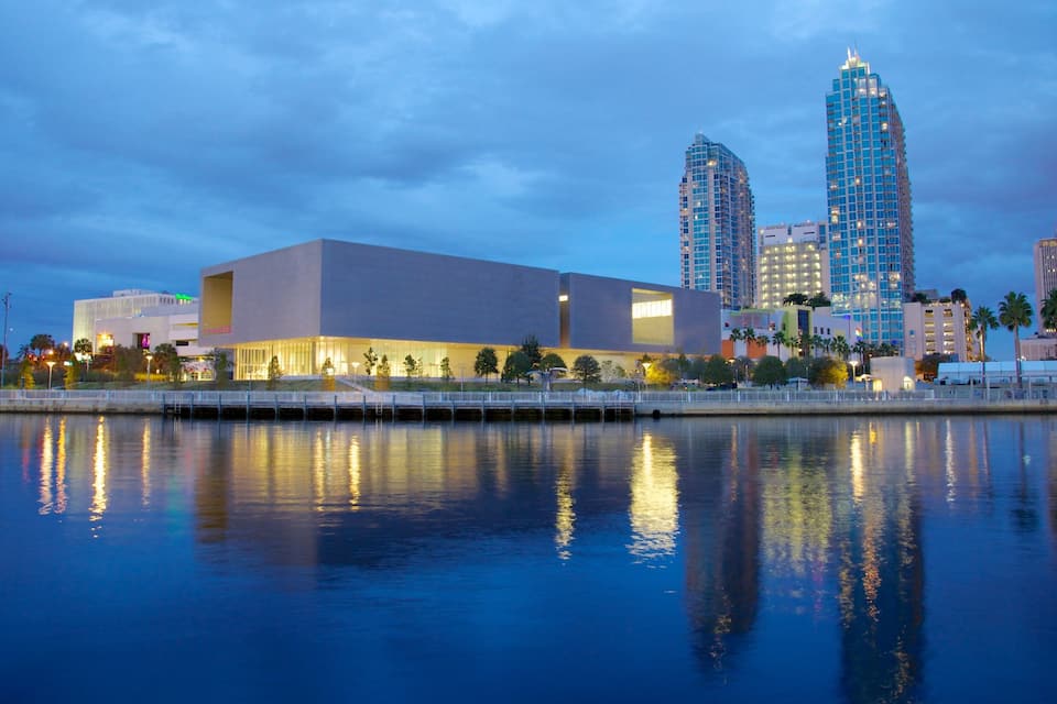 Um edifício moderno com vista para uma orla refletora, cercado por arranha-céus e palmeiras.