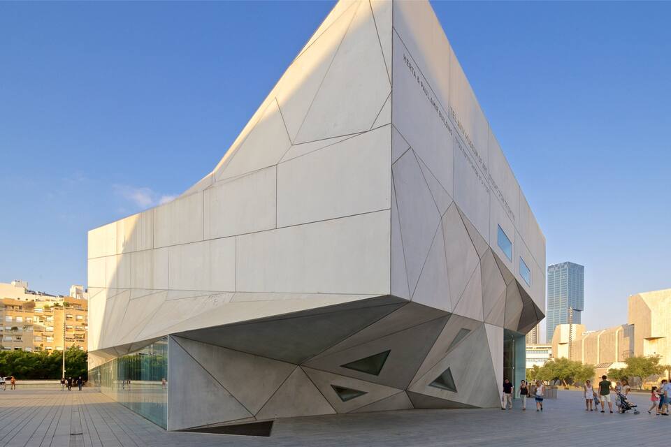 A modern architectural building with a geometric design, surrounded by a plaza with people walking.