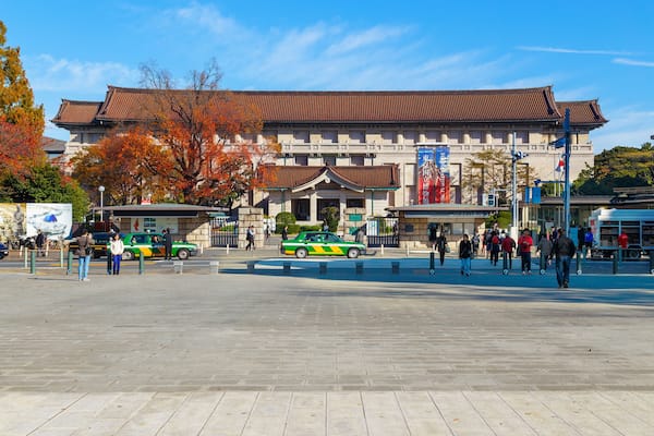 Um edifício tradicional com telhado de azulejos, um carro verde e amarelo, e pessoas caminhando em uma praça.