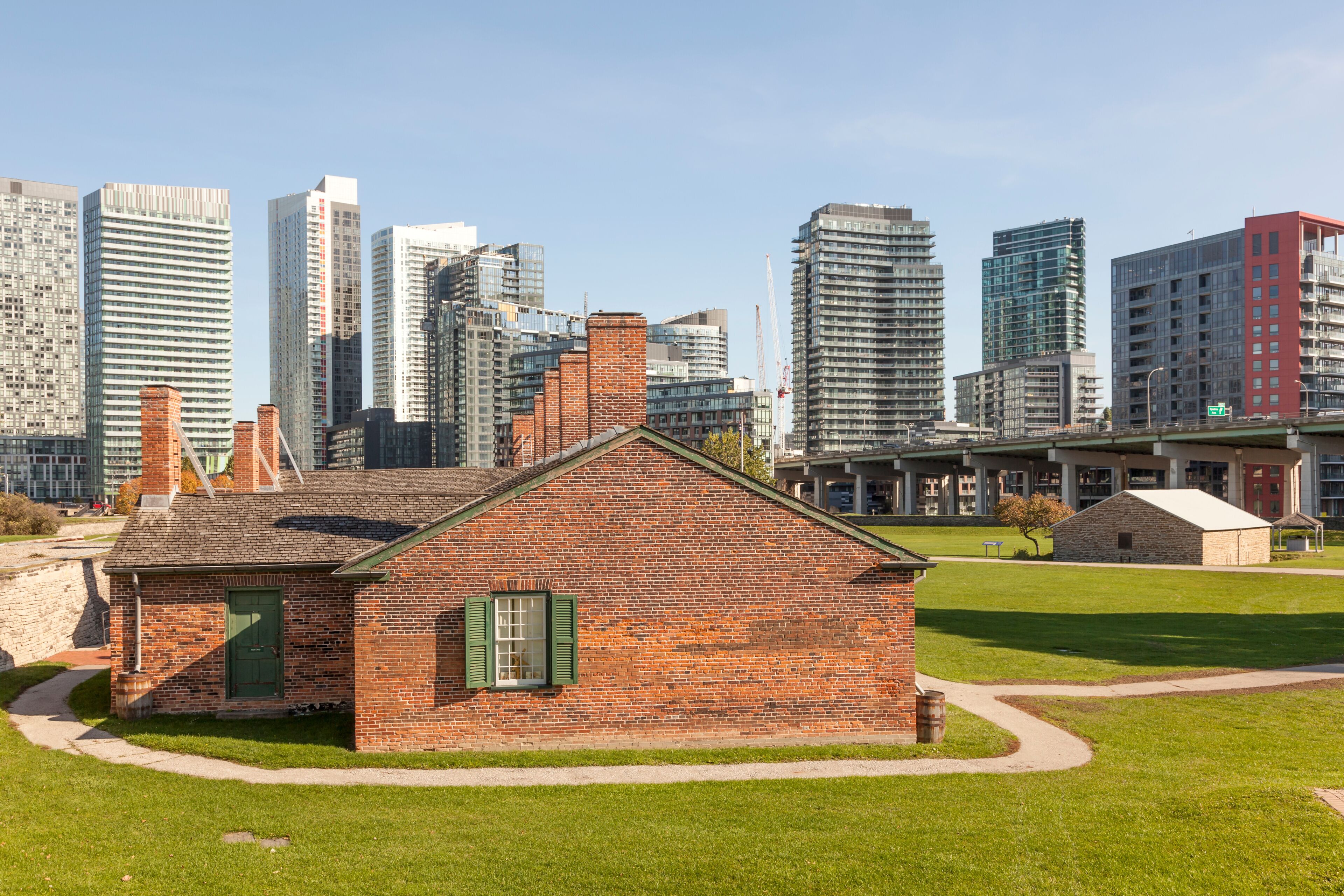 A historic brick building with green shutters, surrounded by a well-maintained lawn, and modern high-rise buildings in the background.