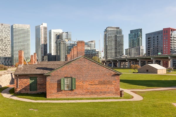 A historic brick building with green shutters, surrounded by a well-maintained lawn, and modern high-rise buildings in the background.