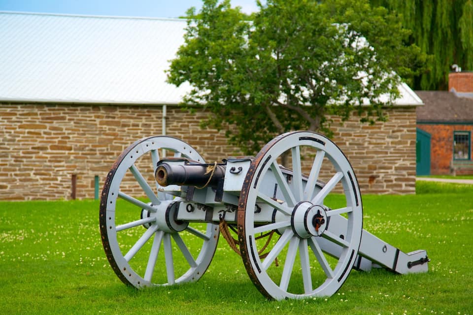Un canon doté de grandes roues blanches se trouve dans un champ herbeux, avec un bâtiment en briques et un arbre en arrière-plan.
