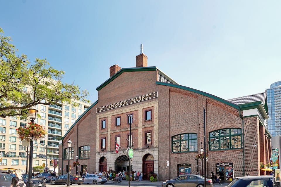 St. Lawrence Market, a historic brick building with large windows and a green roof, surrounded by modern buildings and parked cars.